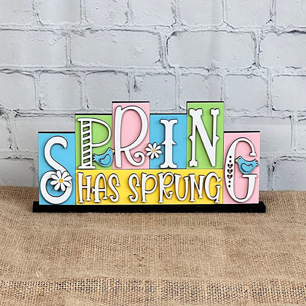 The "Spring Has Sprung" Mini Word Block from Janet's Craft Corner features lively, multicolored letters embellished with patterns, flowers, and a small bird. This charming DIY kit sits on burlap fabric against a white brick wall, bringing a refreshing vibe to any room.