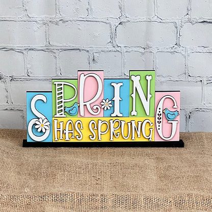 The "Spring Has Sprung" Mini Word Block from Janet's Craft Corner features lively, multicolored letters embellished with patterns, flowers, and a small bird. This charming DIY kit sits on burlap fabric against a white brick wall, bringing a refreshing vibe to any room.