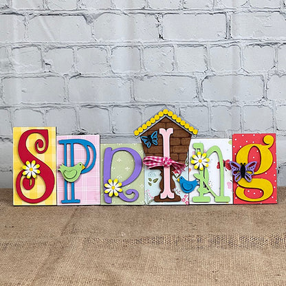 The "Spring Word Block" by Janet's Craft Corner is an ideal addition to home decor, showcasing vivid letters and lively designs of birds, flowers, butterflies, and a quaint house. Displayed against a brick wall on burlap, this delightful piece captures the imaginative charm of a DIY kit with its bright springtime essence.