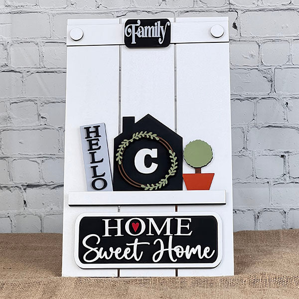 This DIY home decor kit by Janet's Craft Corner, called the Hello Home Monogram Interchangeable Insert, showcases a house adorned with the word "Hello" and a wreath around the letter "C," topped by "Family." A small potted plant sits below, and the base reads "Home Sweet Home" with a heart in the center, set against a brick wall backdrop. It's perfect for craft enthusiasts!