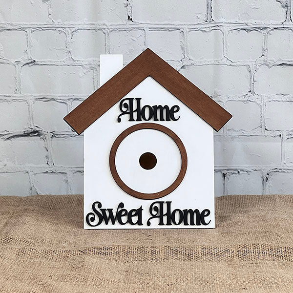 This hand-painted DIY kit from Janet's Craft Corner, called the House Interchangeable Set, includes a charming wooden birdhouse crafted in the shape of a house. It features a brown roof and circular entrance. The words "Home" and "Sweet Home" are elegantly scripted across the white front, making it an ideal addition to any lovely home décor with its backdrop of brick walls.