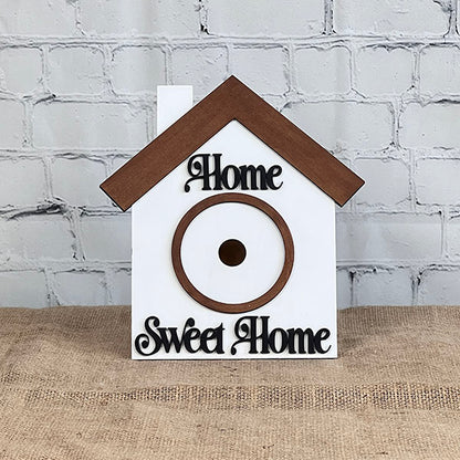 This hand-painted DIY kit from Janet's Craft Corner, called the House Interchangeable Set, includes a charming wooden birdhouse crafted in the shape of a house. It features a brown roof and circular entrance. The words "Home" and "Sweet Home" are elegantly scripted across the white front, making it an ideal addition to any lovely home décor with its backdrop of brick walls.