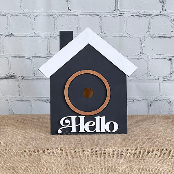 Introducing the House Interchangeable Set from Janet's Craft Corner, a captivating addition to any Home Décor collection. This delightful piece showcases a birdhouse painted in black with a contrasting white roof, set against a brick wall backdrop. Adorned with the welcoming word "Hello" at the bottom in white, it features a circular entrance framed by a wooden ring and rests gracefully on a burlap-covered surface. Ideal for adding charm to any cozy space!