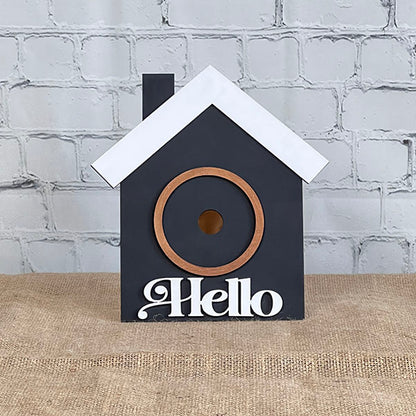 Introducing the House Interchangeable Set from Janet's Craft Corner, a captivating addition to any Home Décor collection. This delightful piece showcases a birdhouse painted in black with a contrasting white roof, set against a brick wall backdrop. Adorned with the welcoming word "Hello" at the bottom in white, it features a circular entrance framed by a wooden ring and rests gracefully on a burlap-covered surface. Ideal for adding charm to any cozy space!