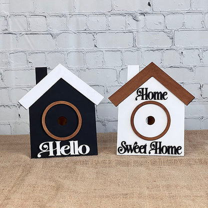 Introducing the House Interchangeable Set by Janet's Craft Corner: two delightful birdhouses perfect for elevating your home décor. The first features a sleek black design with a white roof and a welcoming "Hello." The second offers a cozy white structure complemented by a brown roof and the heartfelt phrase "Home Sweet Home." Displayed against a white brick wall on burlap, these birdhouses capture the spirit of style and creativity.