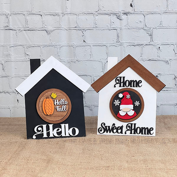 Two enchanting, hand-painted house-shaped signs from Janet's Craft Corner's House Interchangeable Set are displayed on a burlap surface against a white brick wall. The black sign features the phrase "Hello Fall" with an acorn motif, while the white one says "Home Sweet Home" adorned with a Santa face and snowflakes—an exquisite addition to any home décor setup.