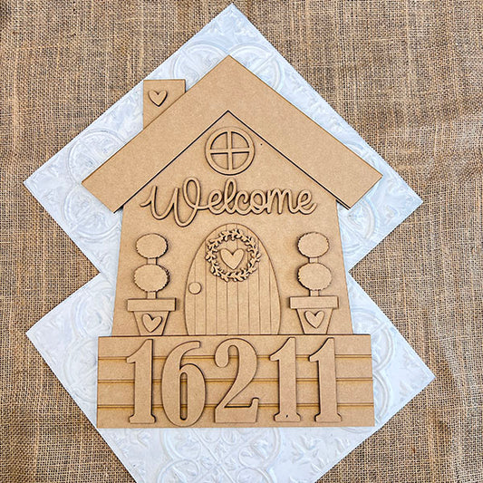 This charming house-shaped wooden plaque by Janet's Craft Corner, ideal for home décor, features a "Welcome" above a round door with a heart wreath. House number "16211" adorns the bottom against a burlap backdrop, making it a perfect addition to your DIY home decor craft kit.