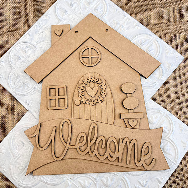 This delightful DIY Kit from Janet's Craft Corner features a wooden house cutout with a rounded door, circular window, and heart-shaped wreath. With "Welcome" at the bottom against a woven-patterned background, it’s perfect as a Welcome Home Door Hanger.