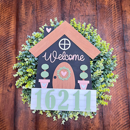 Introducing the House Number Wreath Insert by Janet's Craft Corner—this charming DIY kit includes a decorative wooden house-shaped plaque featuring "Welcome" surrounded by green foliage. Adorned with heart designs on the door, two potted plants, and house number "16211" at the bottom, it makes for a delightful piece of home décor.