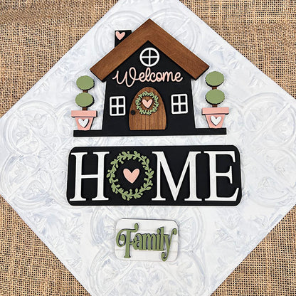 Introducing the "Welcome Home Interchangeable Insert," a DIY home decor kit by Janet's Craft Corner. This decorative insert features a house design with hand-painted lettering, including "Welcome" at the top and "HOME" adorned with a heart-shaped wreath in the "O," followed by the word "Family." The background showcases a charming burlap and white quilted appearance, making it ideal for versatile home decor.