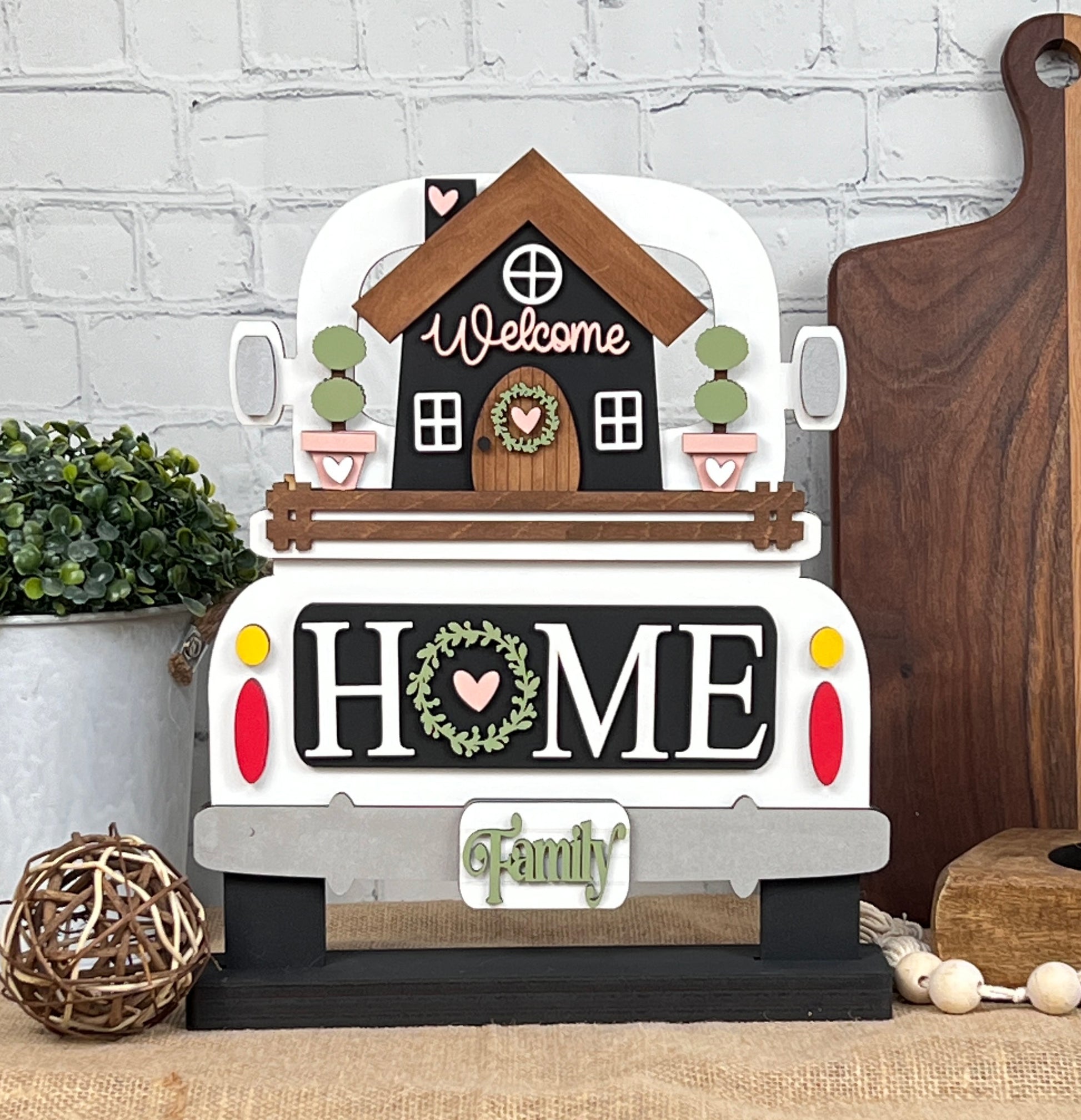 The DIY kit, "Antique Truck Base with Insert" from Janet's Craft Corner, features a decorative sign shaped like an antique truck with a house on top, showcasing "HOME" and "Family" in rustic style along with small plants and hearts. It's set against a brick wall and wooden boards, near a potted plant.