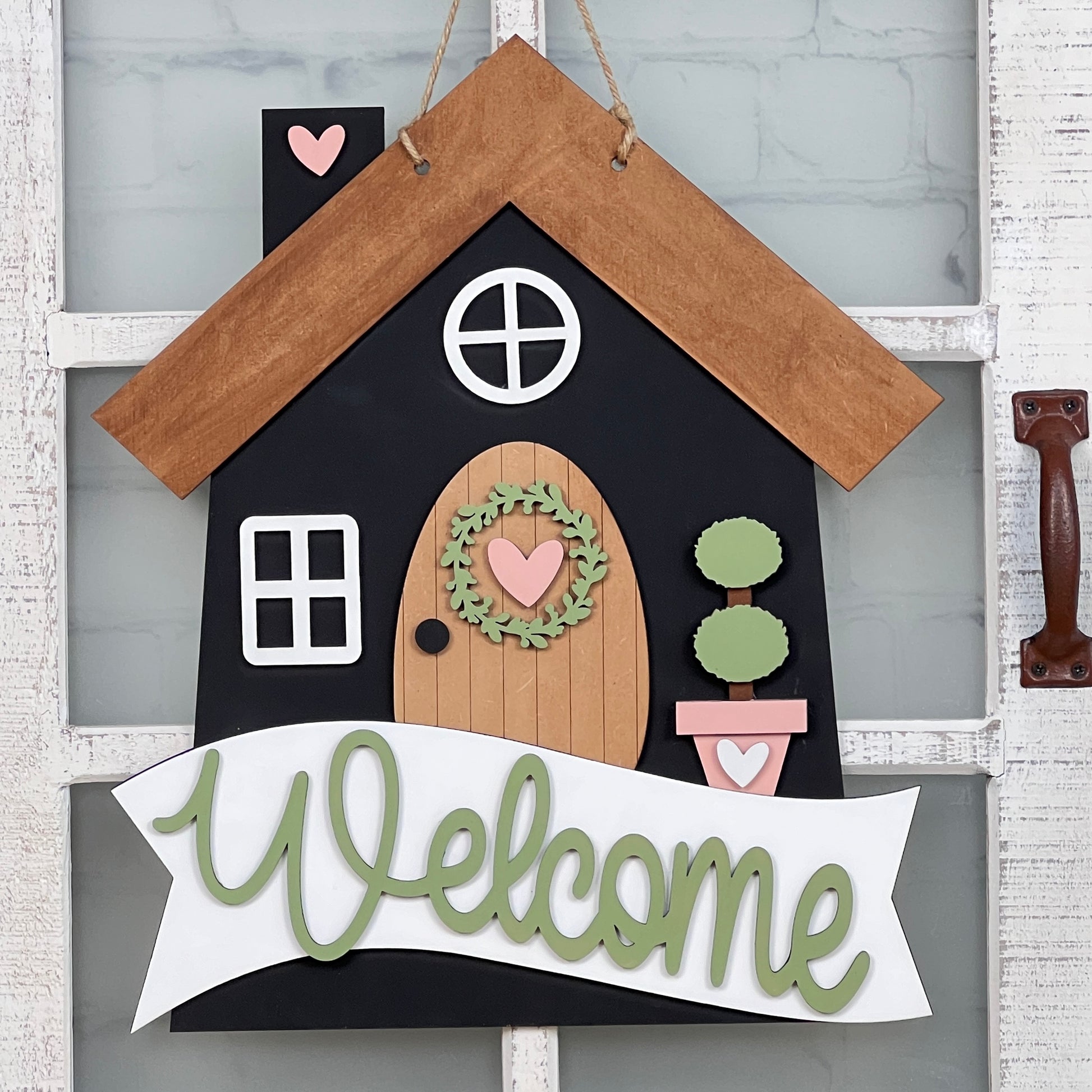 The DIY Kit from Janet's Craft Corner enables you to craft a delightful wooden Welcome Home Door Hanger. With its house shape, the hanger features a brown roof, black walls, and a circular window. It's adorned with a door that has a green wreath, potted plants, and a "Welcome" banner in green letters—making it an ideal addition to your home décor.