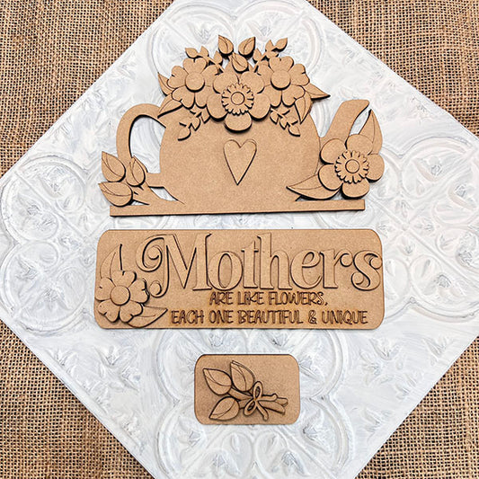 The "Mothers Interchangeable Insert" DIY home decor kit by Janet's Craft Corner includes a wooden teapot cutout adorned with flowers and a heart, along with a sign that says, "Mothers are like flowers, each one beautiful & unique." This delightful craft kit features interchangeable inserts and showcases a small flower bouquet design set against an embossed burlap-textured background.