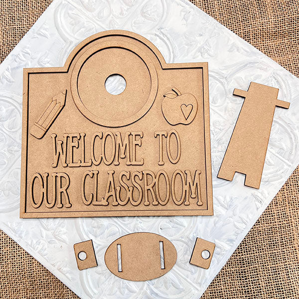 The Welcome to Our Classroom Interchangeable Set by Janet's Craft Corner features wooden cutouts forming a DIY sign with designs like a pencil, apple, and heart. This home decor craft kit includes 1 base and 12 inserts for customizable style.