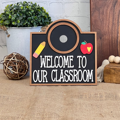 The "Welcome to Our Classroom Interchangeable Set" by Janet's Craft Corner features a delightful classroom décor piece shaped like a camera with a chalkboard center that displays "Welcome to Our Classroom" in white letters. Adorned with a pencil and apple, it effortlessly complements rustic décor, making it an ideal addition to any home setting.