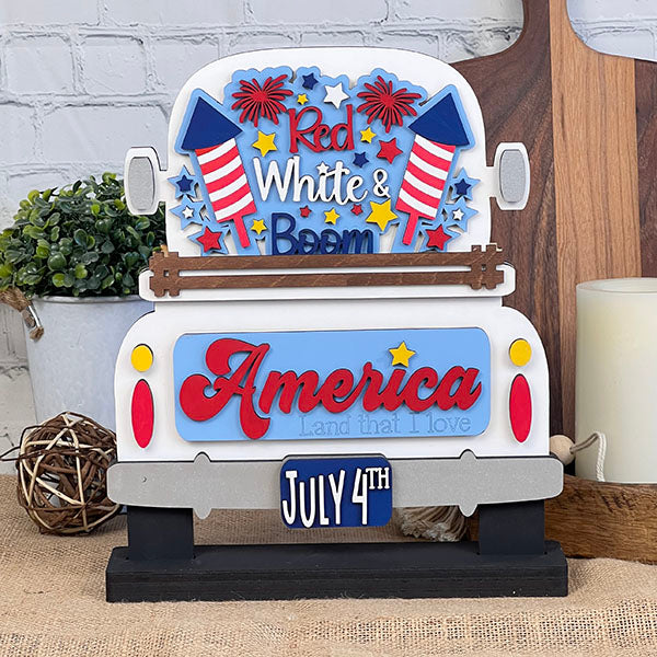 This patriotic wooden decoration by Janet's Craft Corner, the Red White & Boom Interchangeable Insert - Painted, features a white truck filled with fireworks and stars, adorned with "Red White & Boom" on the back. "America" is showcased below alongside a "July 4th" license plate. It makes a perfect addition to home decor set against a rustic backdrop with plants and complementary decor.