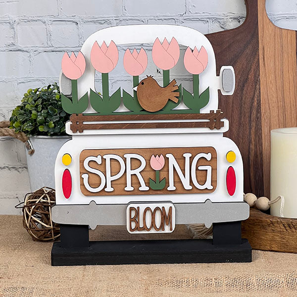 The Spring Tulips Interchangeable Insert from Janet's Craft Corner is a delightful DIY home decor piece. Shaped like the back of a truck, it showcases pink tulips and a brown bird alongside the words "Spring Bloom." Set against a rustic background with a woven mat and plants, it's perfect for adding seasonal flair to your space.