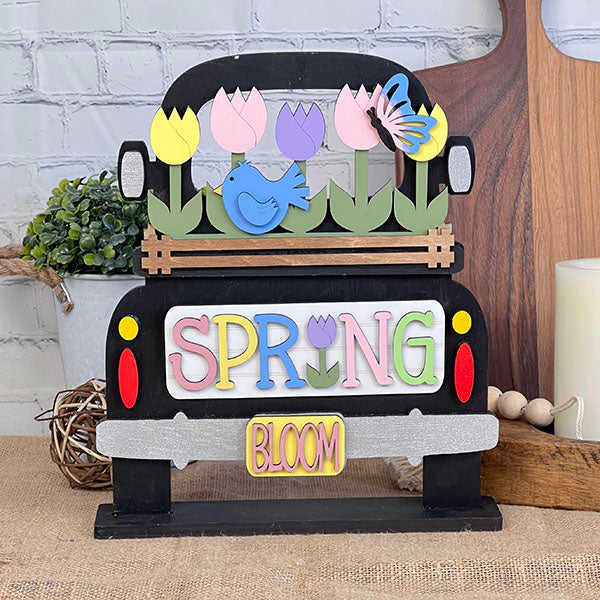 The Spring Tulips Interchangeable Insert from Janet's Craft Corner is a decorative handmade wooden truck adorned with vibrant tulips, a butterfly, and a bird. The word "SPRING" is elegantly displayed on the side with a small "BLOOM" sign beneath it. This charming piece, set against a rustic background, serves as both delightful home decor and an interchangeable insert for various seasons.