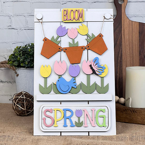 Introducing the Spring Tulips Interchangeable Insert, a delightful DIY home decor kit by Janet's Craft Corner. This charming piece showcases the words "BLOOM" and "SPRING," along with colorful cutouts of tulips, a bluebird, and a butterfly. Designed for home decor enthusiasts, it includes small hanging pots on a rustic white board enhanced with greenery, all available as a hand-painted craft kit.