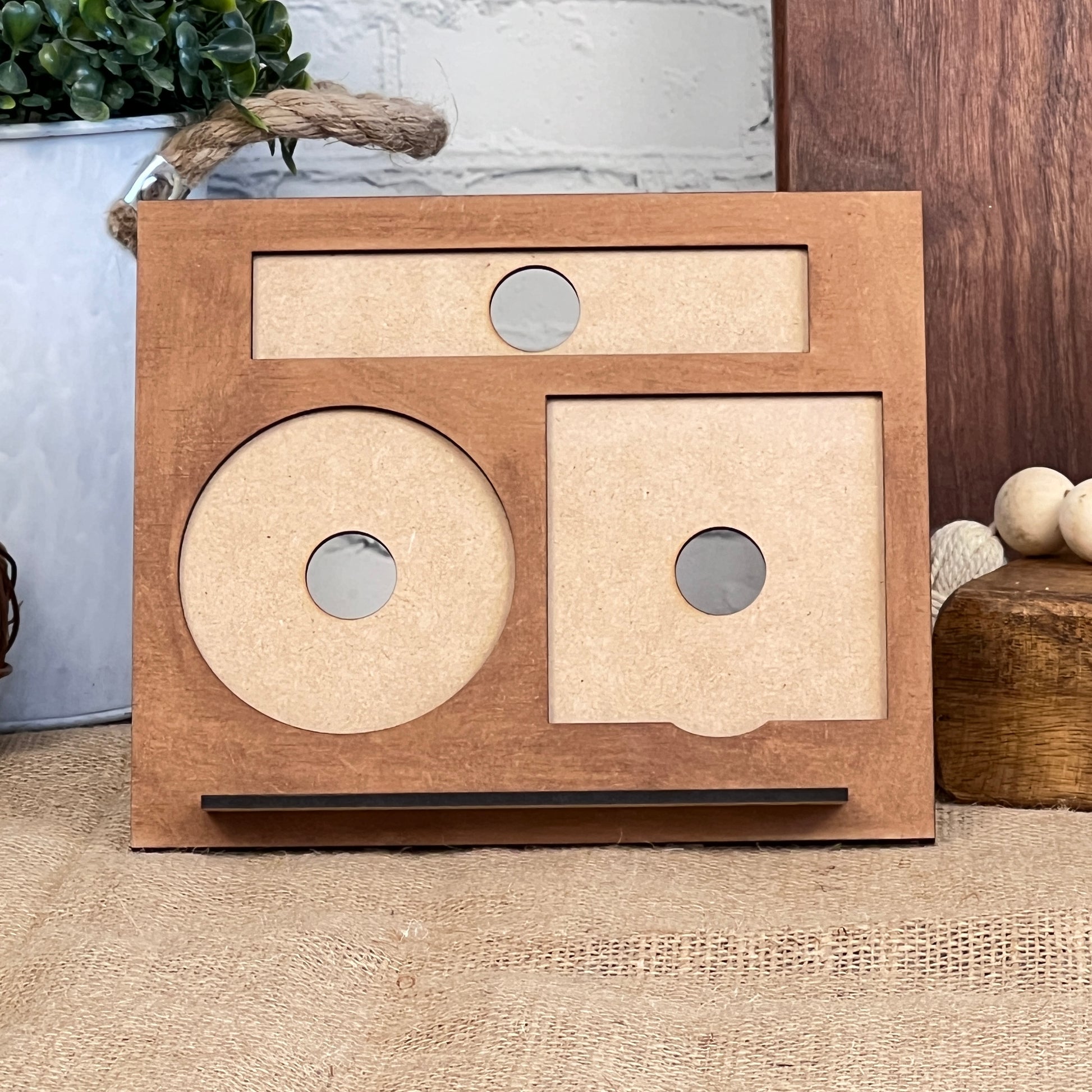 A Desk Post-It Note Holder from the Janet's Craft Corner Interchangeable Set, designed with three sections featuring circular cutouts, leans against a light brick wall. It is surrounded by decorative items like plants and wooden spheres, giving off an interchangeable set vibe. The background includes jute fabric and a wooden block.