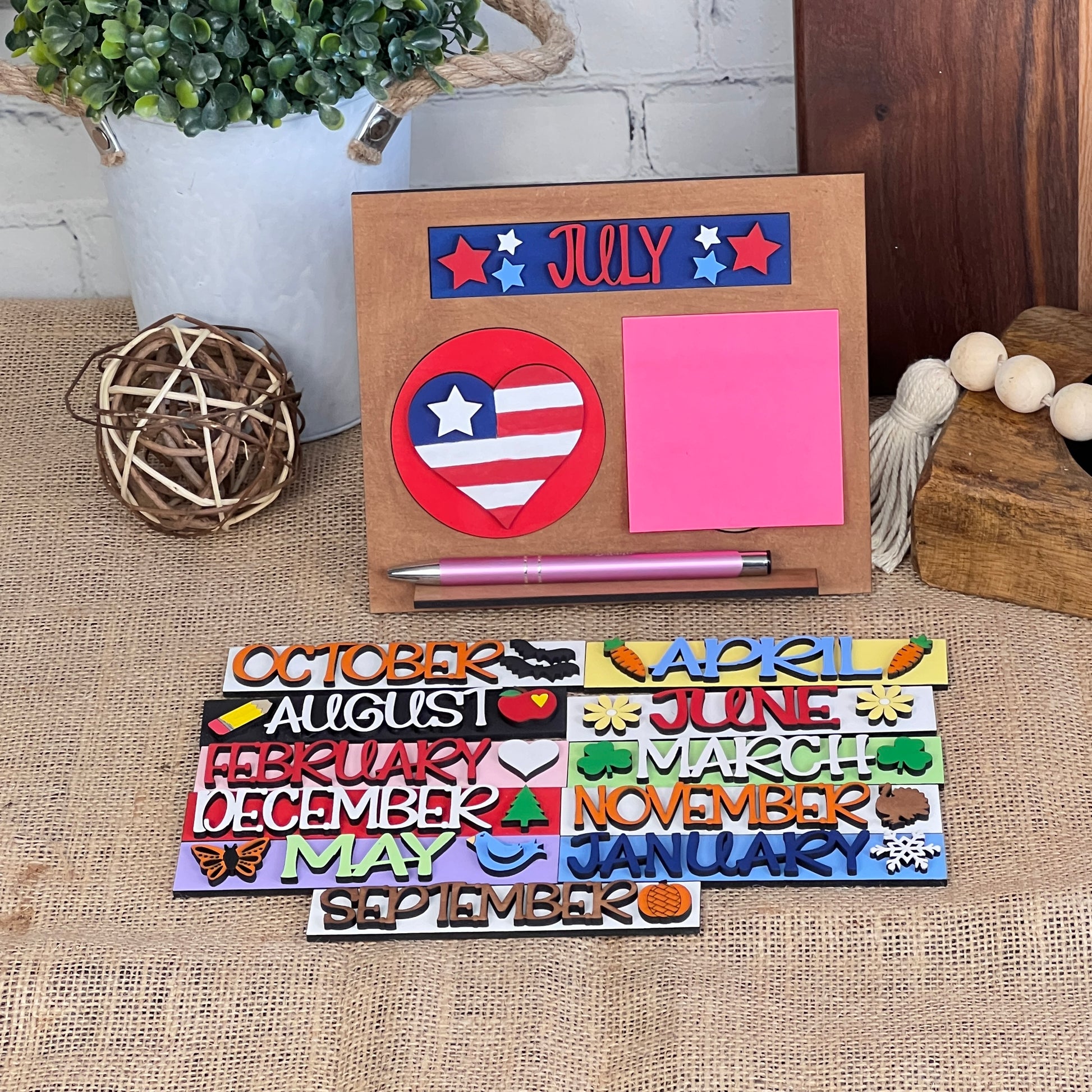 A decorative "Desk Post-It Note Holder Interchangeable Set" from Janet's Craft Corner is highlighted by its wooden design, interchangeable month plaques, pink sticky note, and a heart-shaped USA flag motif. Accompanied by a pink pen, it's displayed on a burlap surface with other rustic decor items.