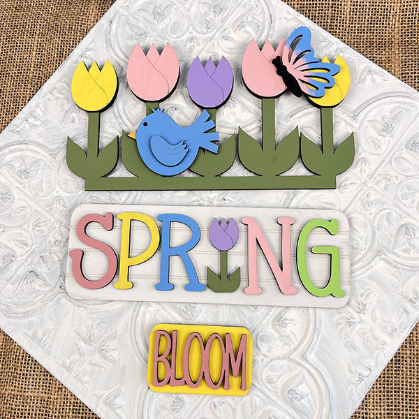 A vibrant wooden art piece from Janet's Craft Corner, designed as an interchangeable insert for home decor, showcases tulips, a blue bird, and a butterfly against a textured background. The words "SPRING" and "BLOOM" are creatively crafted in lively pastel hues. Perfectly suited for adding a splash of seasonal charm to any space!