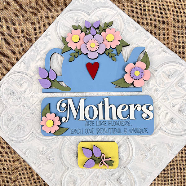 A decorative sign from Janet's Craft Corner, named "Mothers Interchangeable Insert - Painted," features a textured fabric with the phrase "Mothers are like flowers, each one beautiful & unique" in white lettering on a blue background. It is enhanced by a hand-painted teapot decor with pink floral details above and complemented by a small yellow rectangle with purple leaves below.