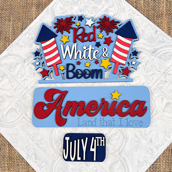 The Red White & Boom Interchangeable Insert - Painted by Janet's Craft Corner features colorful fireworks and stars, celebrating American Pride with the text "Red, White & Boom" at the top, "America, Land that I love" in the middle, and "July 4th" at the bottom. This decorative piece is perfect for enhancing your home decor with its textured background flair.