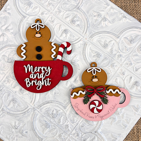 Two gingerbread figures sit cozily in cups on a decorative tile background. The figure on the left holds a candy cane, with the cup displaying "Merry and Bright," making it an ideal piece for holiday decor. The figure on the right is adorned with peppermint and holly, resting in a pink cup featuring the text "Gingerbread Houses & Peppermint Wishes." These charming figures are from Janet's Craft Corner and are part of the Gingerbread Teacup Ornaments collection.