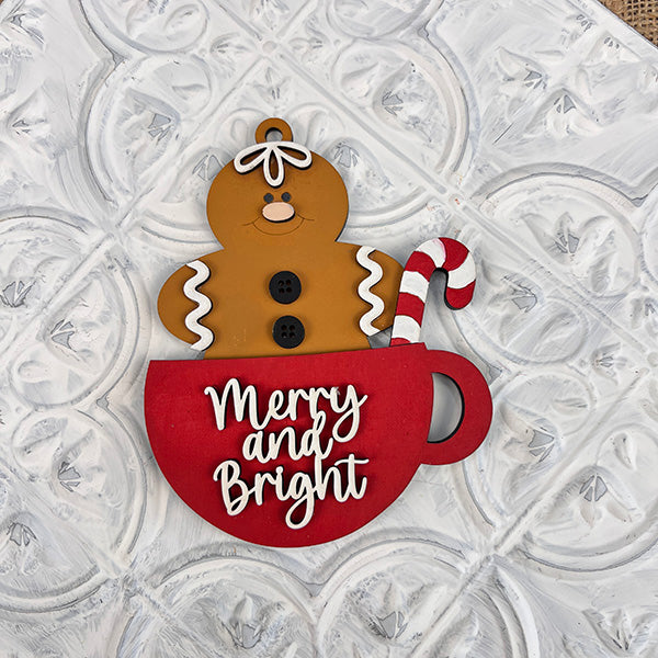 A delightful gingerbread figure holding a candy cane playfully emerges from a red cup adorned with the text "Merry and Bright," making it an ideal holiday decoration. This charming display can be featured in your collection of Gingerbread Teacup Ornaments - Painted from Janet's Craft Corner, infusing your celebrations with whimsical cheer.