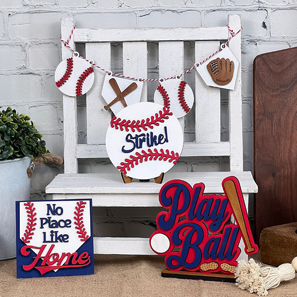 A wooden chair displays a Baseball Tiered Tray Set from Janet's Craft Corner, featuring a baseball-themed home décor with a DIY kit charm. A garland decorated with baseballs and gloves gracefully drapes at the top, while signs reading "Strike!", "No Place Like Home," and "Play Ball" enhance the setup, evoking the essence of a delightful baseball tiered tray display.