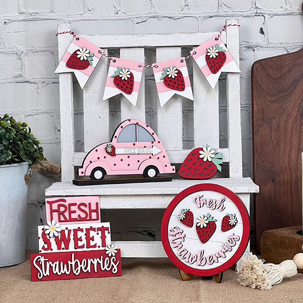 The Strawberry Tiered Tray Set by Janet's Craft Corner creates a delightful display of home décor, featuring a pink car cutout adorned with strawberry designs, a charming banner of strawberries and daisies, and signs proclaiming "Fresh Sweet Strawberries." A white wooden bench acts as the backdrop, enhanced by the charm of a potted plant.