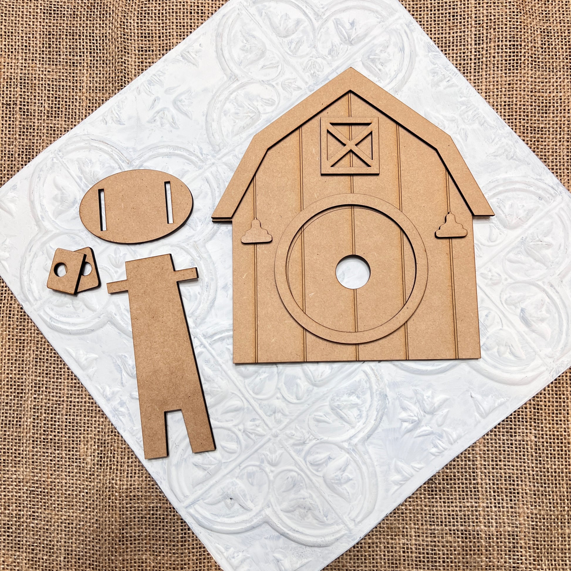 Neatly arranged on a textured white tile, Janet's Craft Corner's Barn Interchangeable Set includes a barn shape with a circular opening and an oval-headed figure, plus small cutout accessories. The hand-painted DIY kit is set against a rustic burlap background.