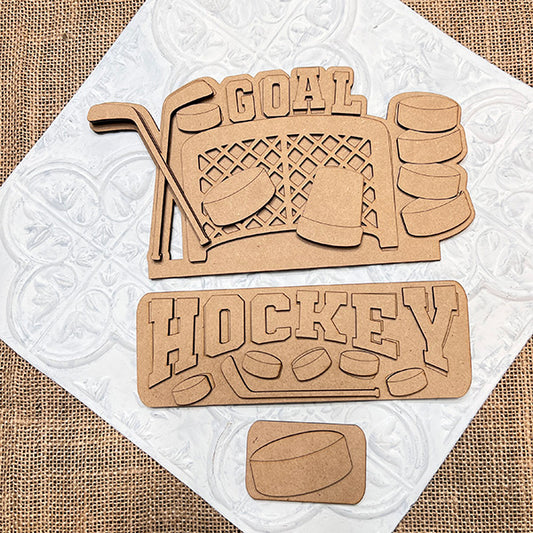 The Hockey Interchangeable Insert DIY home decor kit by Janet's Craft Corner features wooden cutouts with hockey-themed designs arranged on a textured surface. Included in the kit are cutouts of crossed hockey sticks, a goal net, pucks, and the words "GOAL" and "HOCKEY," making it ideal for creating interchangeable home decorations.