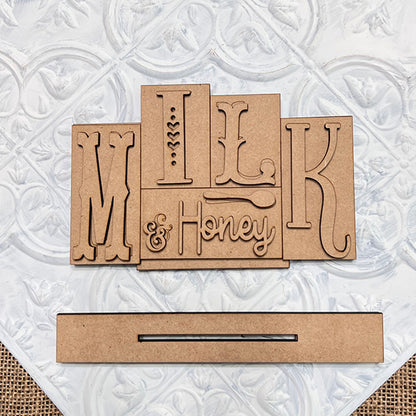 The Milk & Honey Mini Word Block by Janet's Craft Corner is a DIY seasonal home decor craft kit. It features wooden cutout letters on an ornate white background with a rectangular wooden slot for customization.