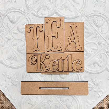 The Tea Kettle Mini Word Block Shelf Sitter from Janet's Craft Corner is a DIY decor kit. It features "TEA Kettle" in decorative font on a textured white background, with an unfinished look and a slot for easy crafting, perfect for tea lovers.