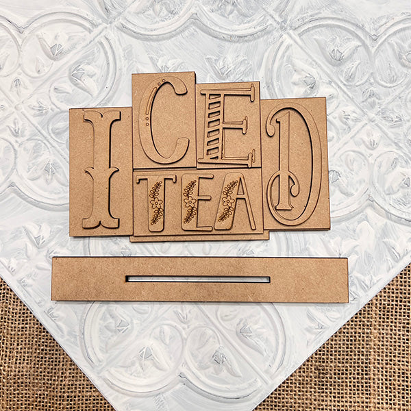 The Iced Tea Mini Word Block by Janet's Craft Corner features overlapping wooden letters spelling "Iced Tea" on a textured white and burlap base, perfect for home décor. A separate wooden strip is included, ideal for DIY seasonal home decor projects.