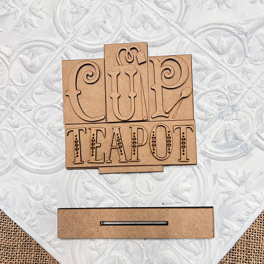 Janet's Craft Corner's Cup-Teapot Mini Word Block Shelf Sitter features decorative wooden cutouts of "CUP" and "TEAPOT" on a textured white surface, ideal for DIY home décor. A slot-shaped cutout enhances its charm.