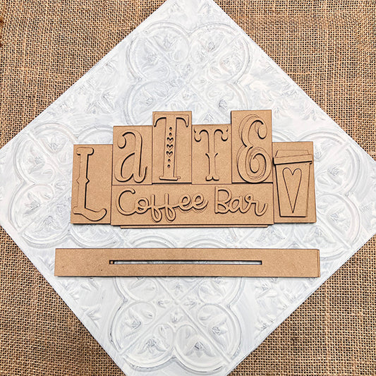 The Latte Mini Word Block from Janet's Craft Corner, a DIY seasonal home decor kit, features "Latte Coffee Bar" spelled out on white tile. A small coffee cup with a heart adds charm to this burlap-surfaced décor piece.