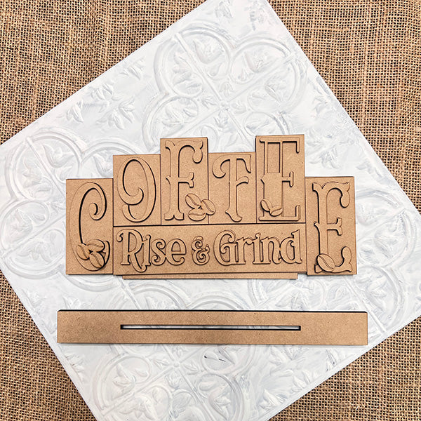 The "Coffee Mini Word Block" by Janet's Craft Corner, features a wooden cutout sign reading "COFFEE Rise & Grind" with coffee bean designs. This DIY craft kit adds charming coffee-themed decor to any space, perfectly displayed on a decorative white square atop burlap fabric.