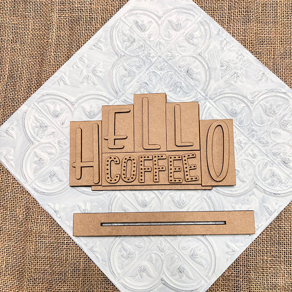 The "HELLO COFFEE" wooden cutouts from Janet's Craft Corner's DIY Kit, ideal for decor lovers, are displayed on a white textured square with a burlap background. A separate thin rectangular piece with a slit is included for assembling your own Hello Coffee Mini Word Block.