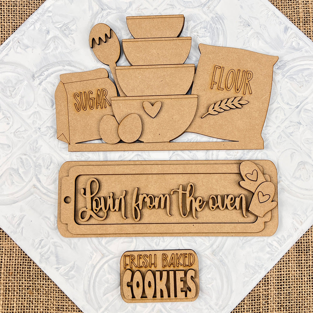 Introducing the "Lovin from the Oven Interchangeable Insert" DIY home decor kit by Janet's Craft Corner. This charming set features hand-painted wooden kitchen-themed cutouts including bowls, eggs, sugar, and flour bags on a textured background. Two delightful plaques read "Lovin' from the oven" with hearts and "Fresh baked cookies." The kit provides interchangeable inserts for a personalized decor touch that’s ideal for any kitchen space.