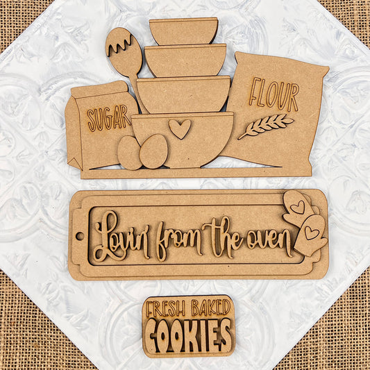 Introducing the "Lovin from the Oven Interchangeable Insert" DIY home decor kit by Janet's Craft Corner. This charming set features hand-painted wooden kitchen-themed cutouts including bowls, eggs, sugar, and flour bags on a textured background. Two delightful plaques read "Lovin' from the oven" with hearts and "Fresh baked cookies." The kit provides interchangeable inserts for a personalized decor touch that’s ideal for any kitchen space.