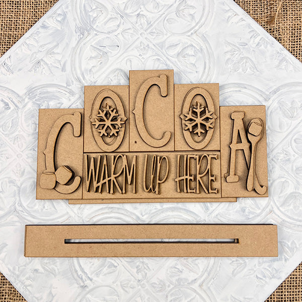 The Cocoa Mini Word Block by Janet's Craft Corner features "COCOA WARM UP HERE" with snowflakes, a mitten, and a mug on a textured white background. This hand-painted wooden sign is perfect for cozy home décor.