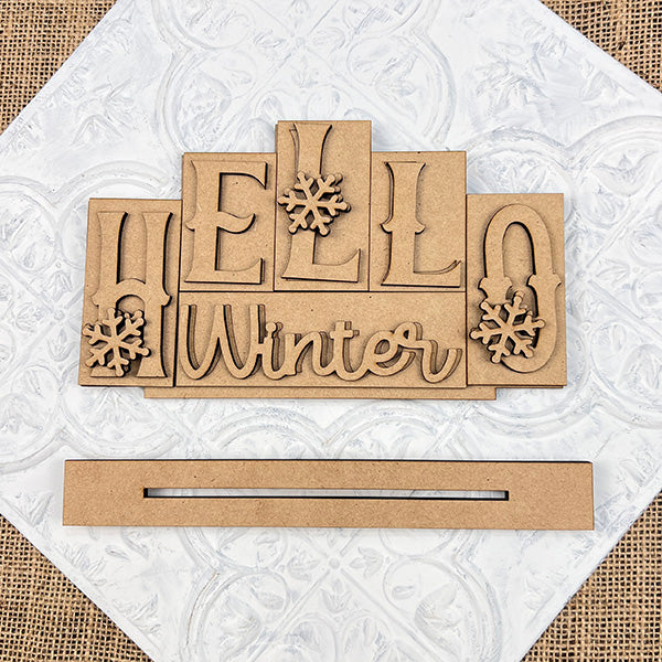 The Hello Winter Mini Word Block Shelf Sitter by Janet's Craft Corner features hand-painted wooden letters with snowflake details on "O" and "E." It's set against a textured white background, with a separate piece below featuring a horizontal slit, ideal for DIY crafts or seasonal home décor.