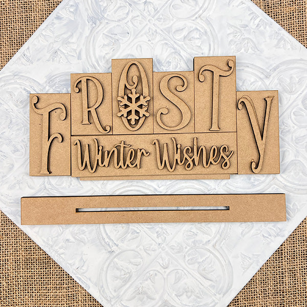 The Frosty Mini Word Block Shelf Sitter by Janet's Craft Corner features "Frosty Winter Wishes" with a snowflake-themed "O," set on a textured white and brown background, ideal for seasonal home decor.