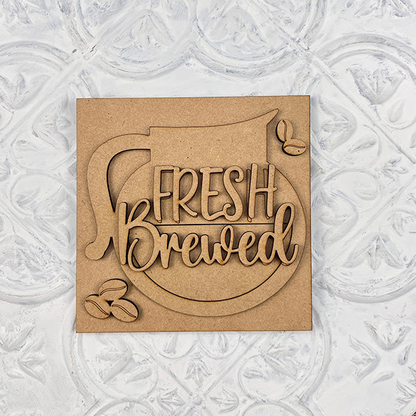 The Coffee Swappable Squares DIY craft kit by Janet's Craft Corner features a wooden coffee pot cutout with "Fresh Brewed" and four coffee beans on a textured white background—perfect seasonal home decor for coffee lovers.