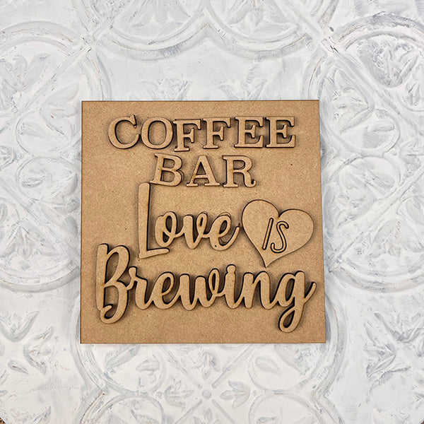 A wooden sign with layered text reads "COFFEE BAR Love is Brewing," with "is" in a heart, on a white textured background—perfect for Janet's Craft Corner Coffee Swappable Squares DIY seasonal home decor craft kit.