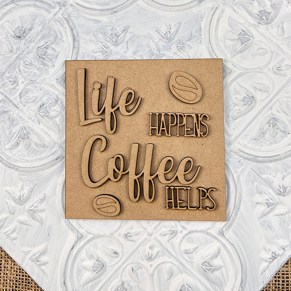A wooden sign on a textured white background reads “Life happens, coffee helps,” surrounded by coffee bean designs—the perfect inspiration for your next Coffee Swappable Squares DIY kit from Janet's Craft Corner.