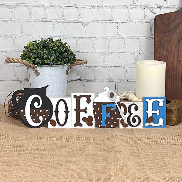 The Coffee Word Block from Janet's Craft Corner spells out "COFFEE," with each letter designed in distinct, coffee-themed styles. Ideal for home décor, it is displayed on a burlap surface set against a white brick wall, complemented by a small potted plant and candle.